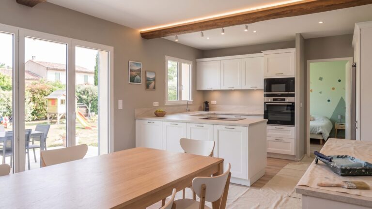 Cuisine blanche ouverte sur un séjour aux murs gris, avec table en bois et poutre apparente