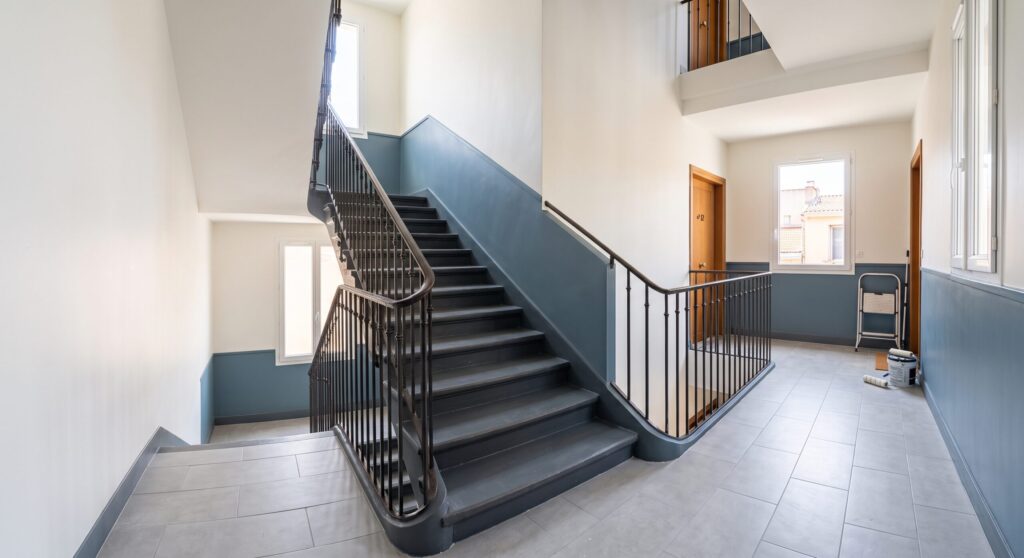 Cage d’escalier avec rampe métallique noire, soubassement bleu et matériel sur le palier