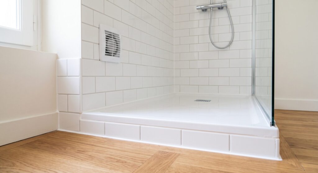 Receveur de douche blanc, carrelage mural clair et grille d’aération, près d’un sol d’aspect bois