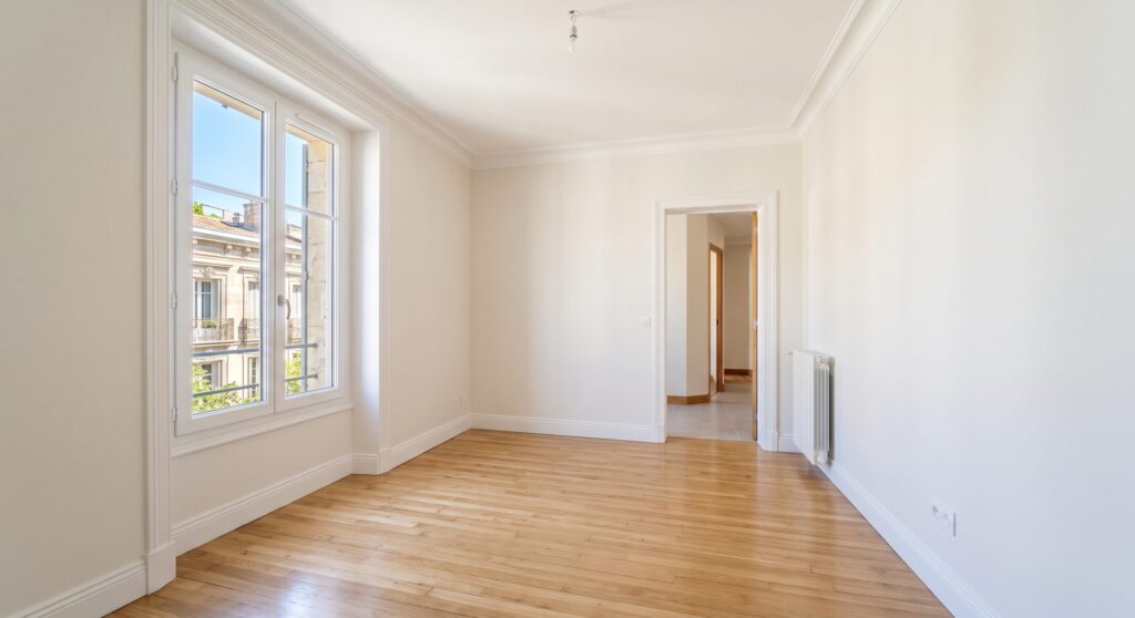 Séjour vide aux murs et plafonds blancs, avec parquet et fenêtres ouvertes sur un balcon